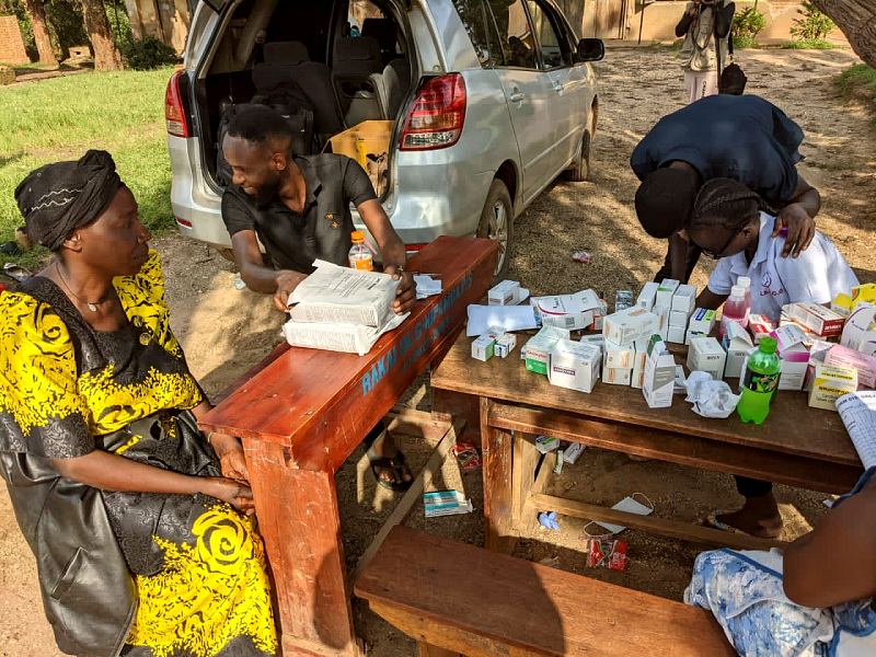 Nurses sort medicines at a wooden desk beside a car with its boot open, under a large tree